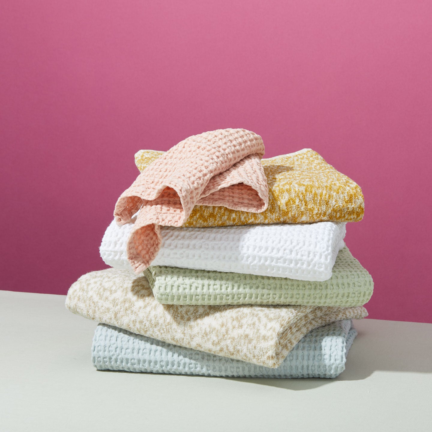 Group of towels in various colors, patterns and textures stacked in front of a pink background.