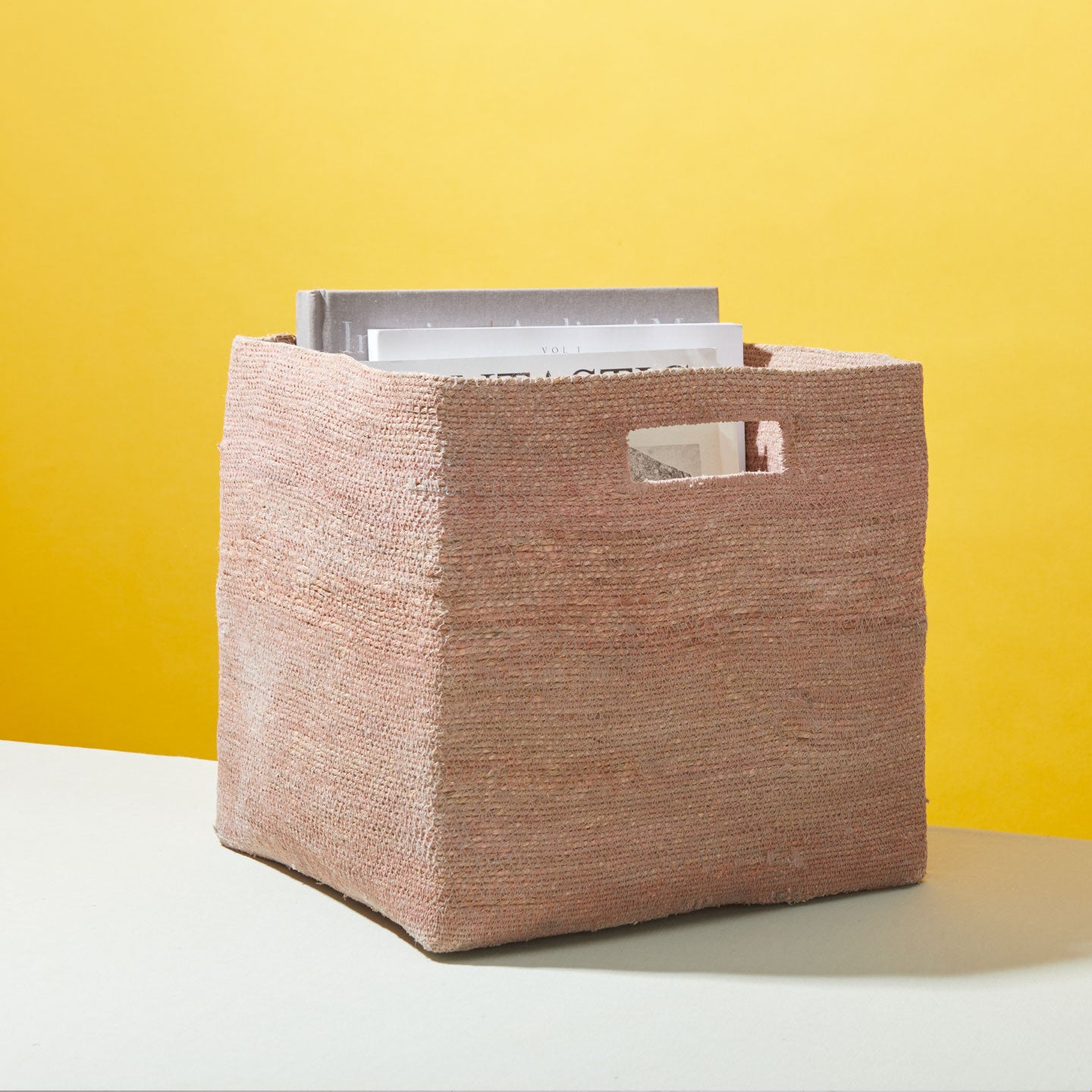 A square, woven basket with handles containing books is placed on a surface against a yellow background.
