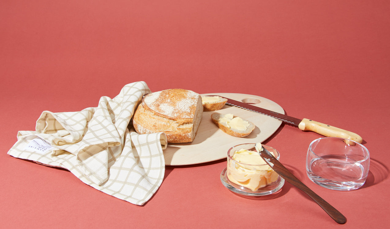 sourdough bread on cutting board with bread knife, dishtowel, butter keeper, and butter spreader