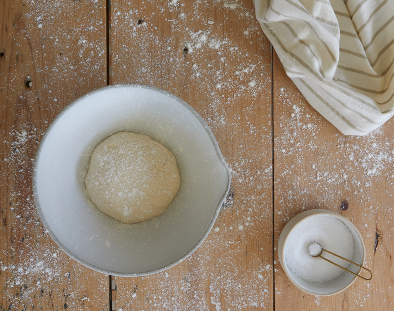 Ball of dough in a bowl on a wooden surface with flour and a towel.