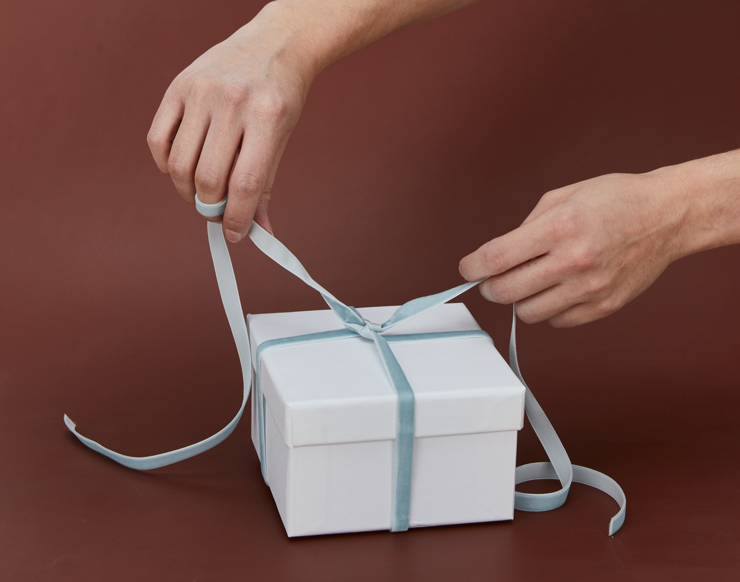 Person tying a blue ribbon on a white gift box against a brown background