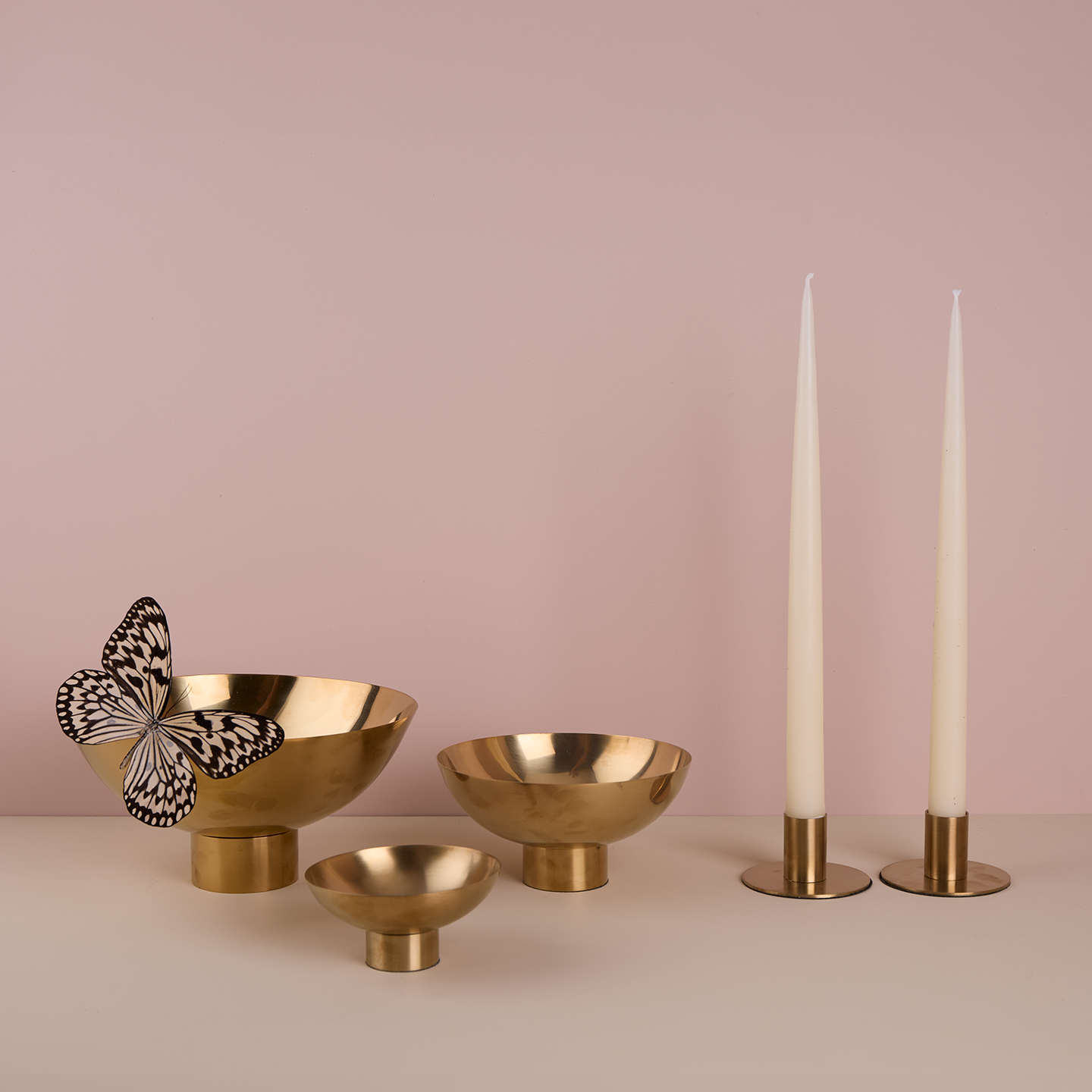 A trio of footed bowls and pair of candle holders, all in Brass, with white taper candles. Styled with a butterfly.