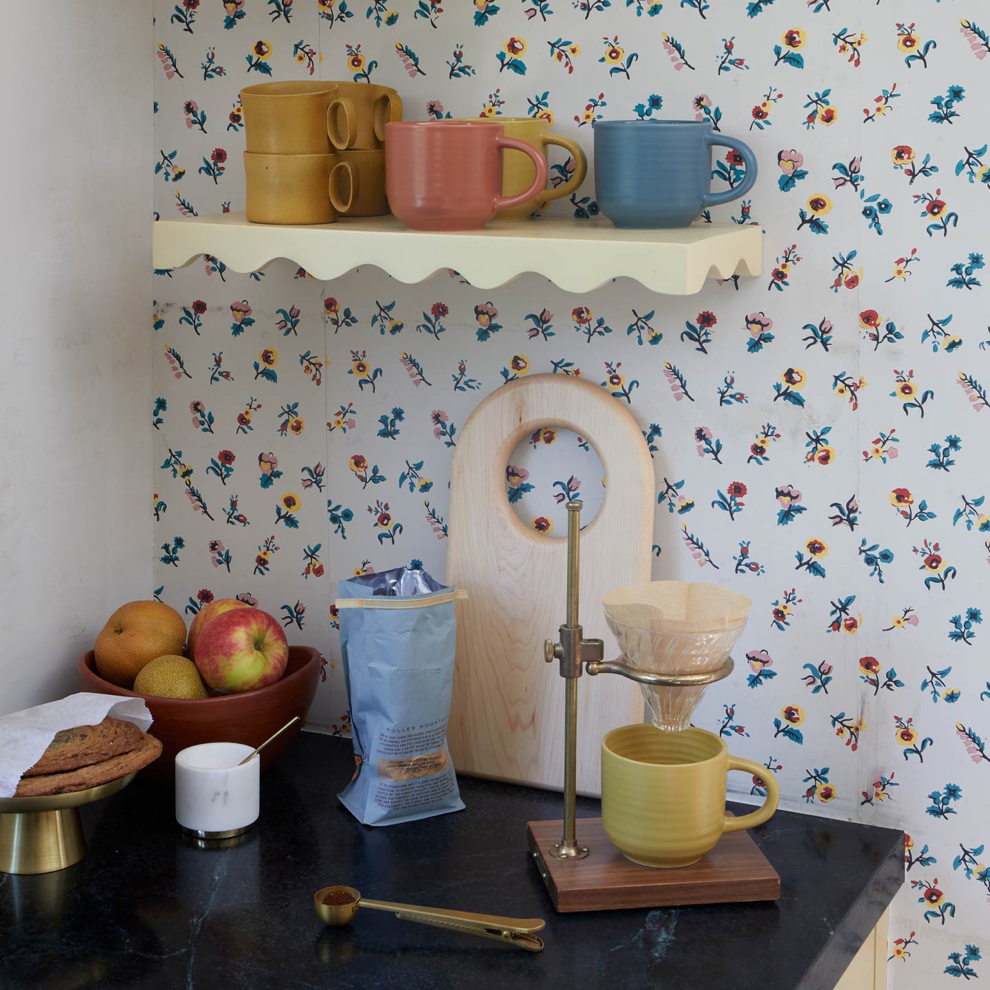 Coffee setup in kitchen with mugs, shelf, coffee, bowl of apples and floral background.