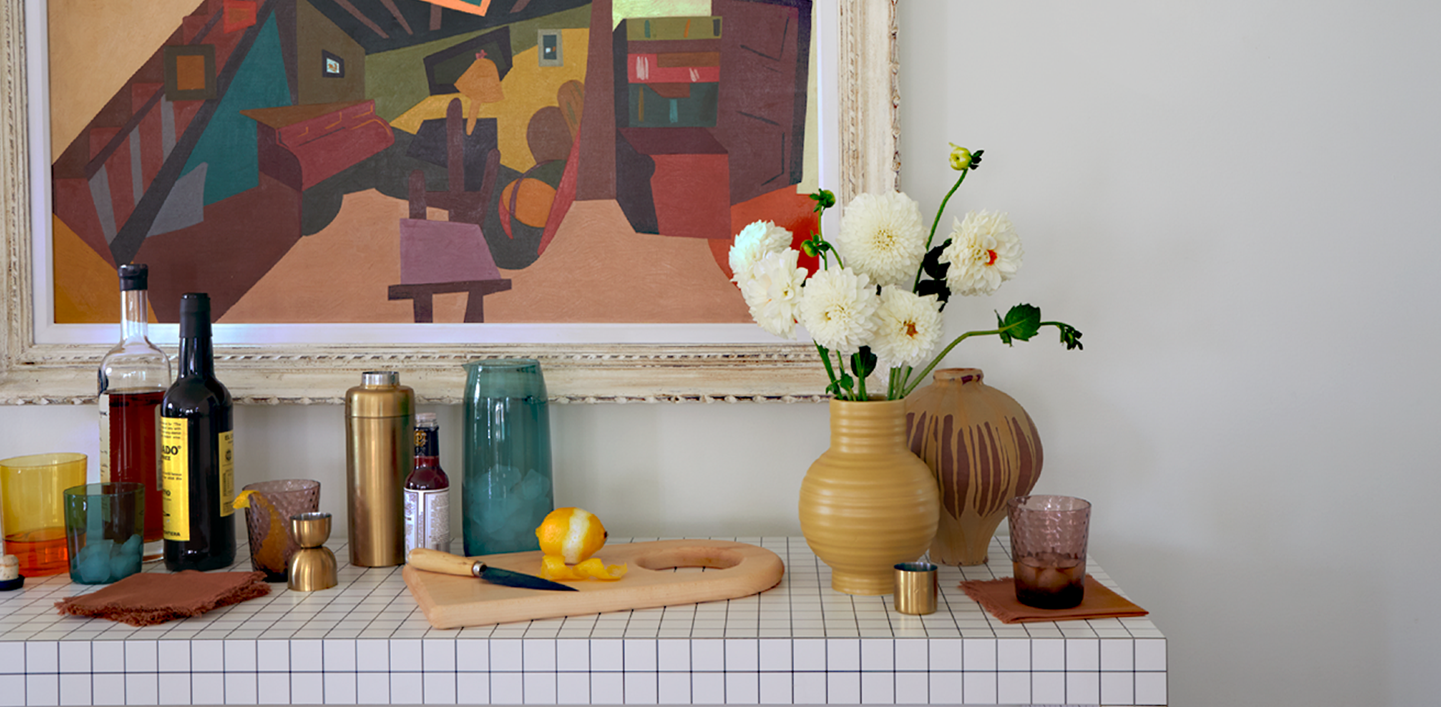 A table with a checkered tablecloth, on top sits various barware, and flowers in a Essential ceramic vase. Behind hangs a large piece of abstract artwork