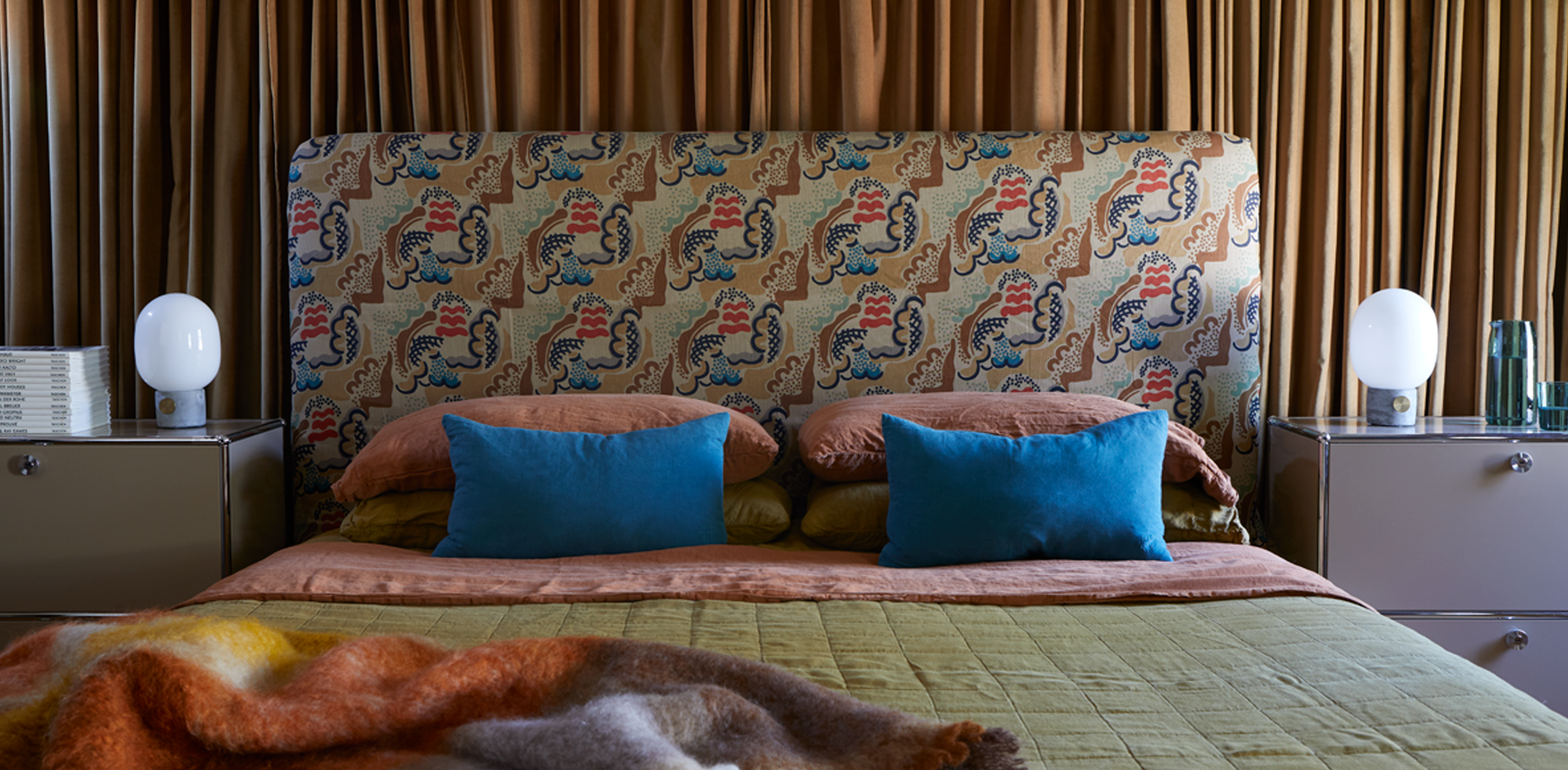 A bedroom with linen pillows in various colors, with quilt and mohair throw, end tables flank the bed with lamps and a glass pitcher with drinking glass