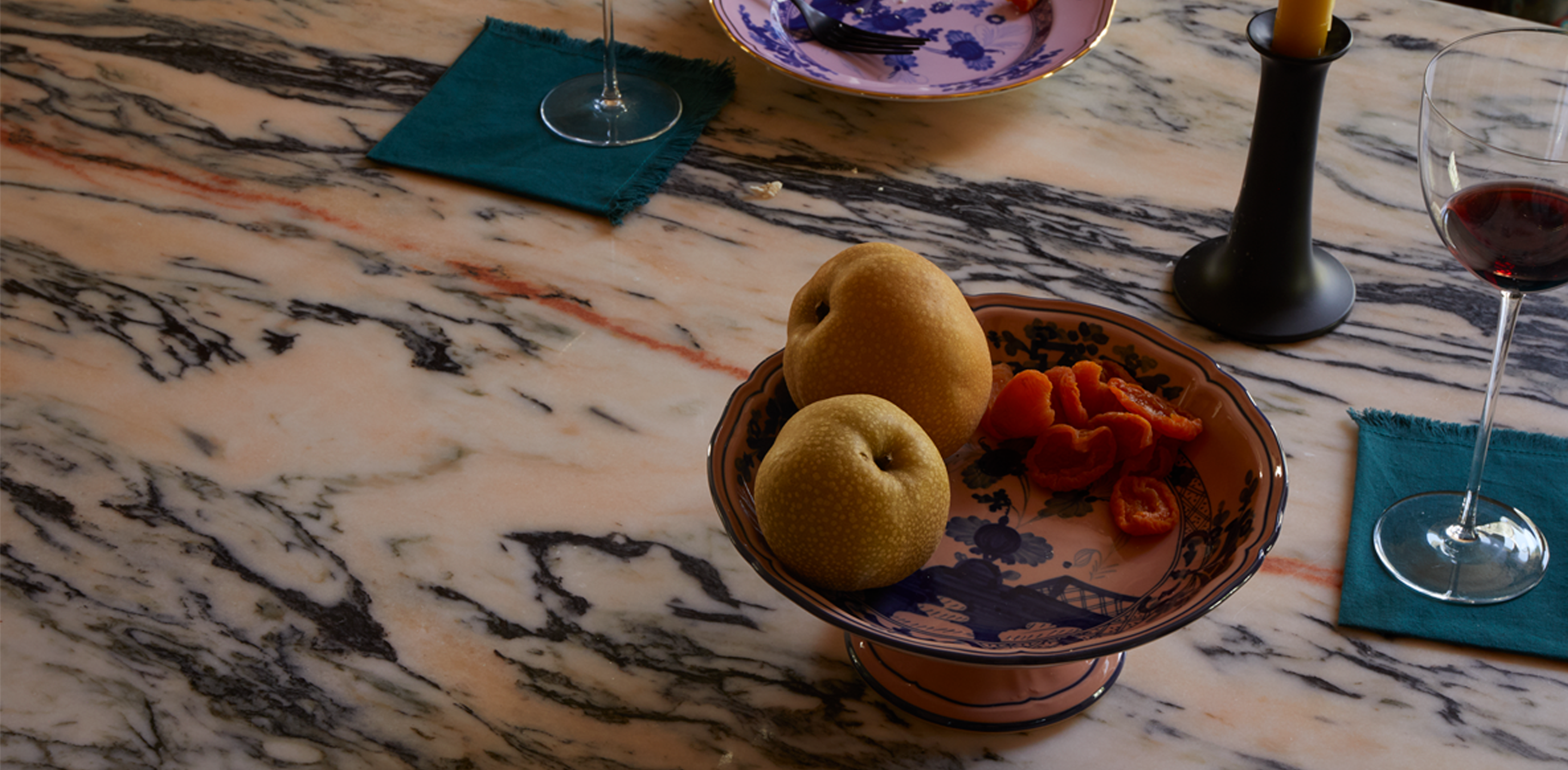 A marble tabletop with a footed fruitbowl filled with pears and citrus, to the sides sit wine glasses on napkins, and taper candle in holder and a dessert plate are placed in the background