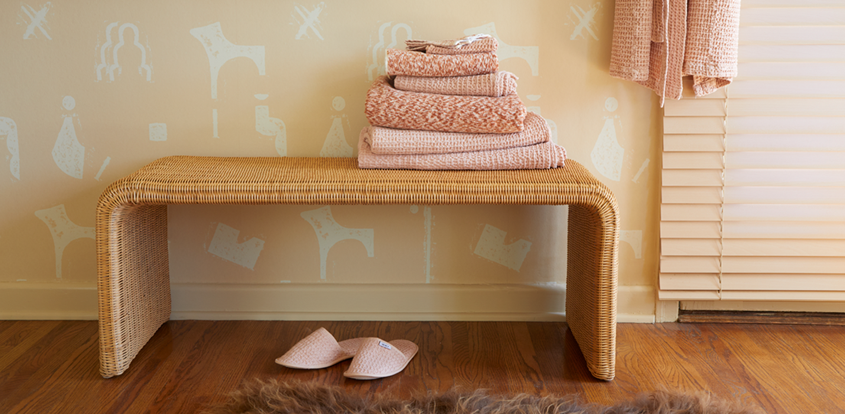 A bathroom with patterned wall paper, wicker bench, and a stack of blush towels, a pair of slippers and fur rug sit below, and a blush robe hangs above