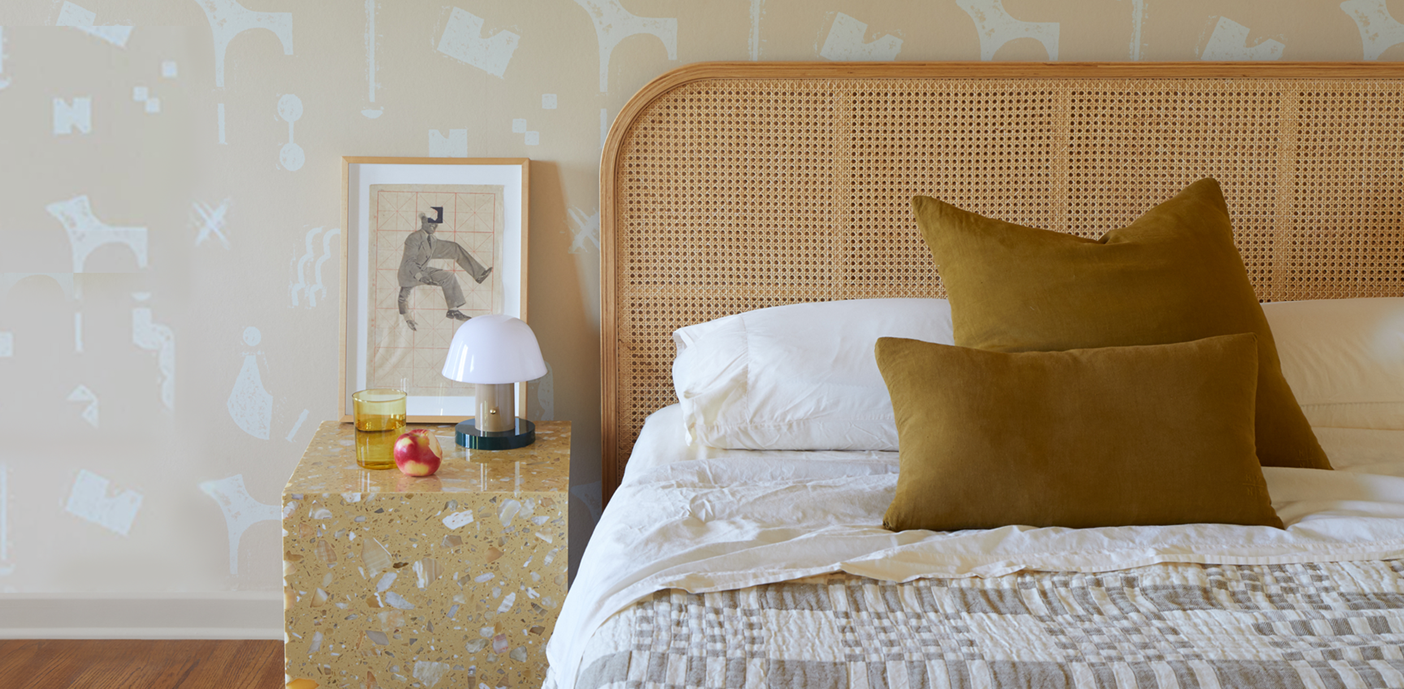 A bed with pillows and a plaid blanked sits next to a marble night stand with a lanp, artwork, and an apple