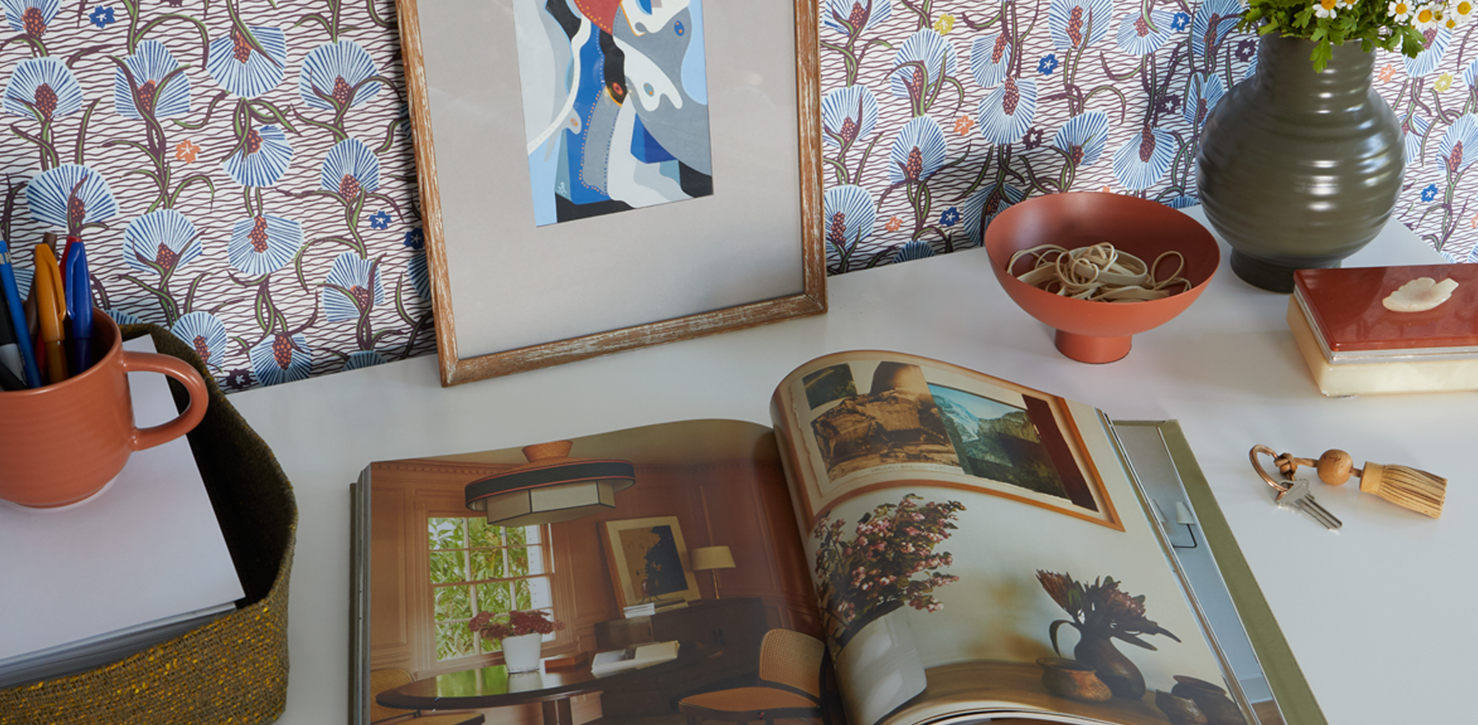 An office table with on open book, a basket of paper and writing utensils. To the right sits a footed bowl containing rubber bands, a large essential vase and keys sit nearby