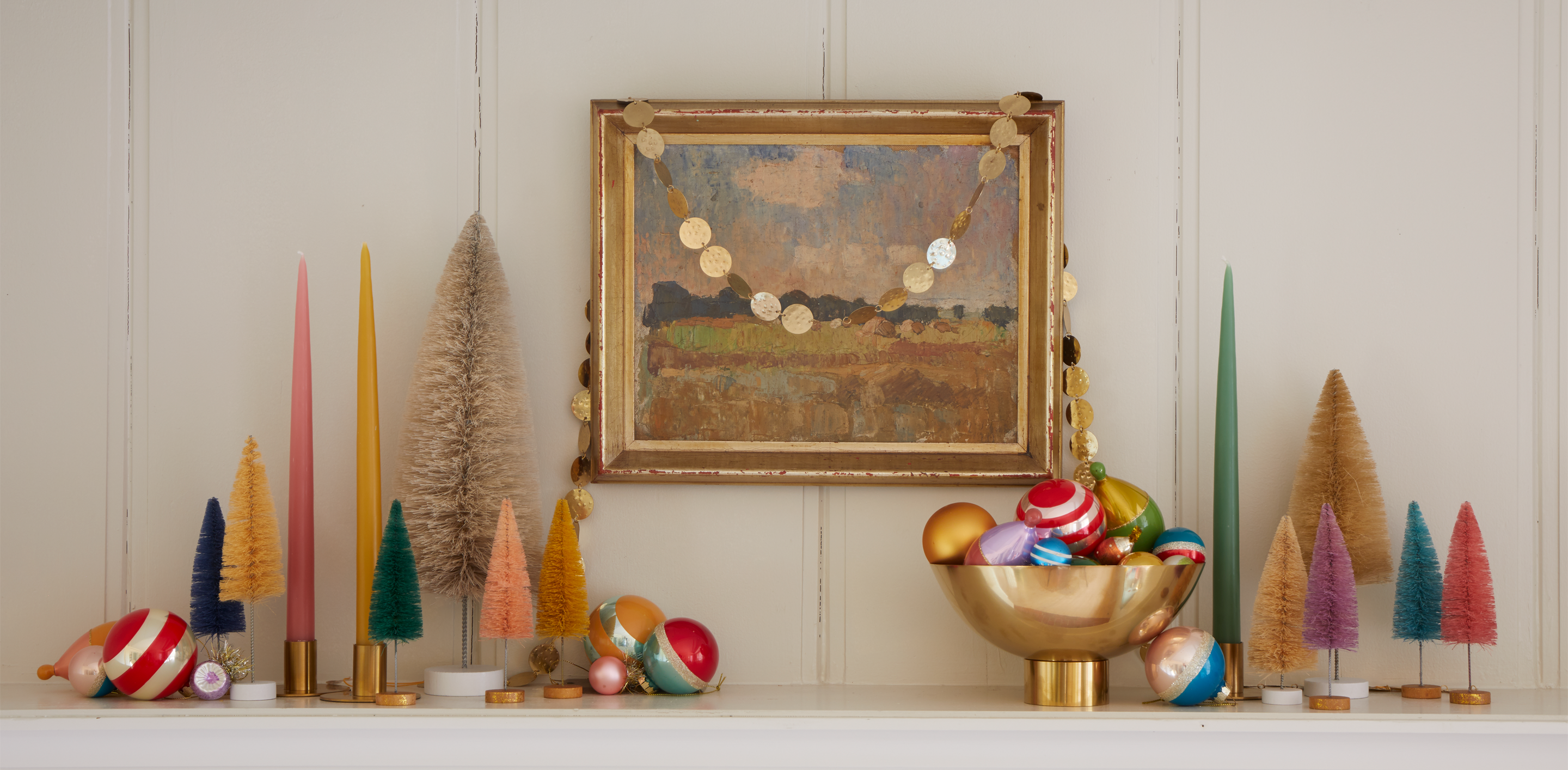 Mantel decorated with bottlebrush trees, taper candles, and a brass footed vase filled with holiday ornaments behind sits framed artwork with a gold garland hung on the frame.