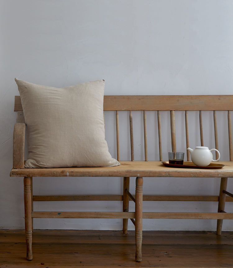 A wooden bench with a beige cushion on one end and a tray with a white teapot and a glass cup on the other against a plain white wall.