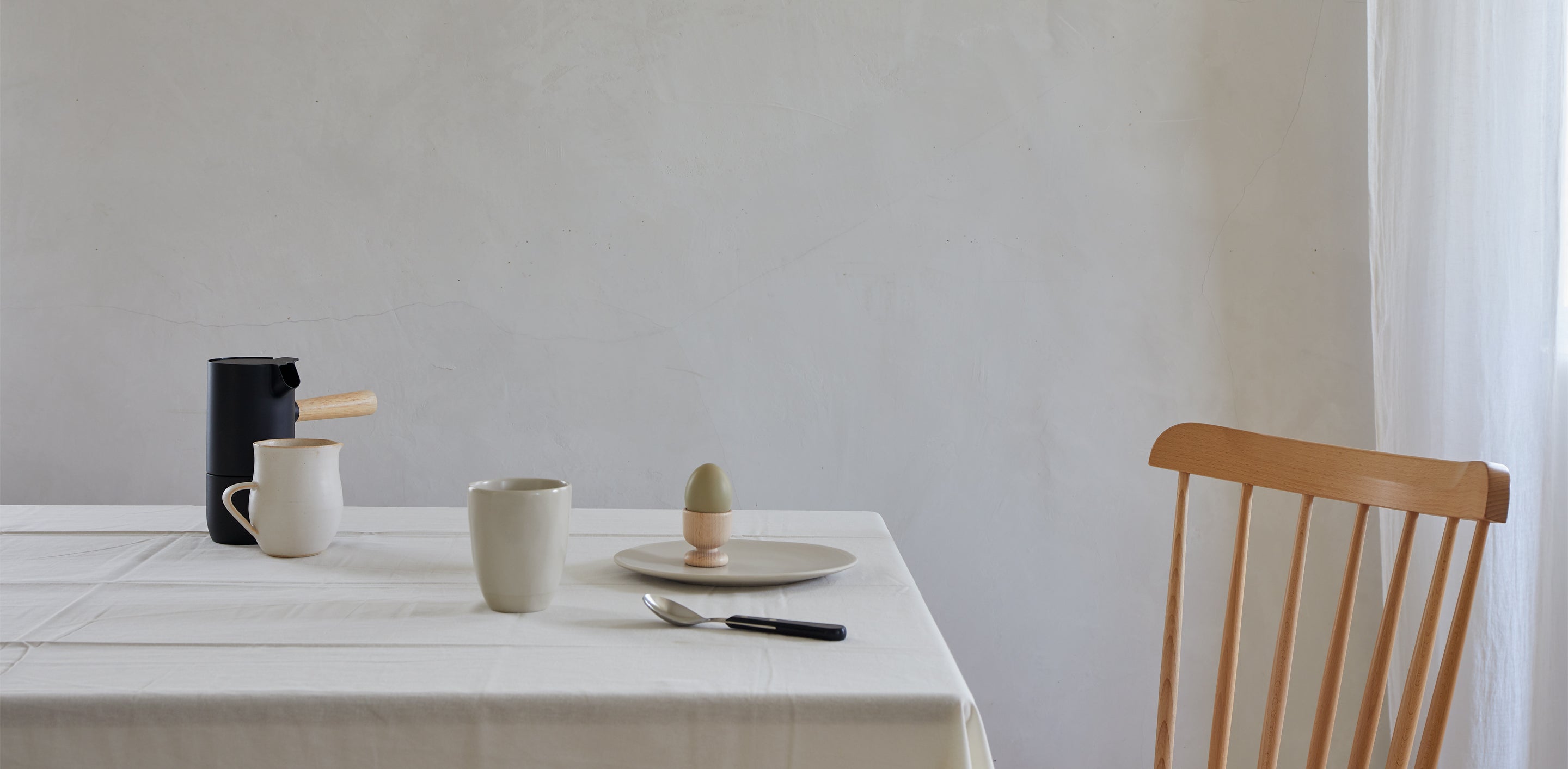 A minimalistic dining setup with a white tablecloth, a wooden chair, a black coffee pot, a white mug, a plate with an egg in an egg cup, and a knife and spoon on the table.