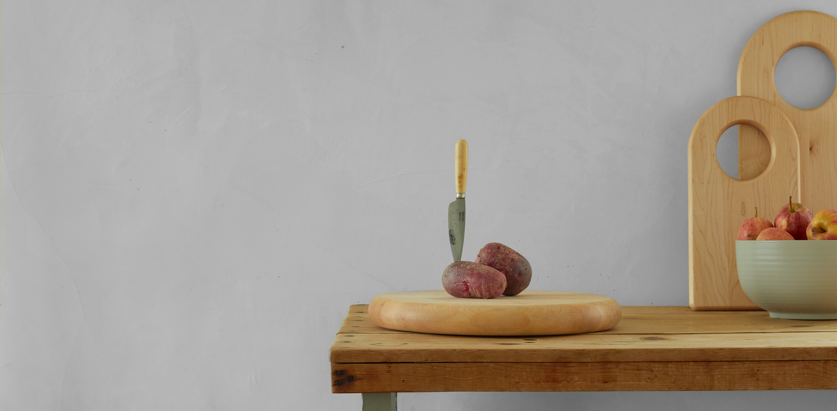 A knife is stuck upright in a potato on a wooden cutting board on a kitchen table. A bowl of apples and two wooden boards are in the background.