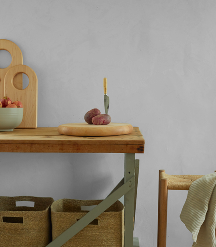 A knife is stuck upright in a potato on a wooden cutting board on a kitchen table. A bowl of apples and two wooden boards are in the background. Baskets are below the table.