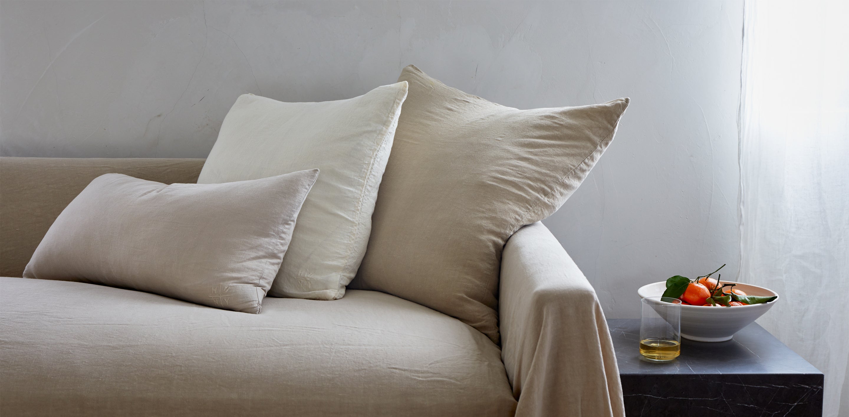 A beige sofa with three pillows sits next to a dark end table holding a bowl of oranges and a glass with a golden liquid.