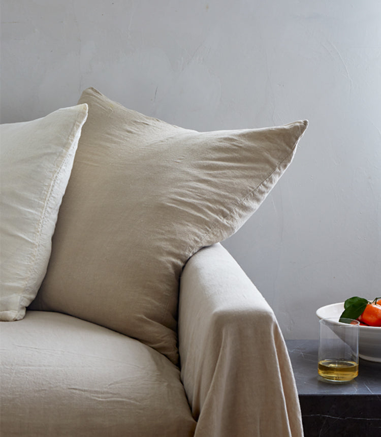 A beige sofa with two large beige pillows is next to a dark side table holding a white bowl with a drink and orange fruit.