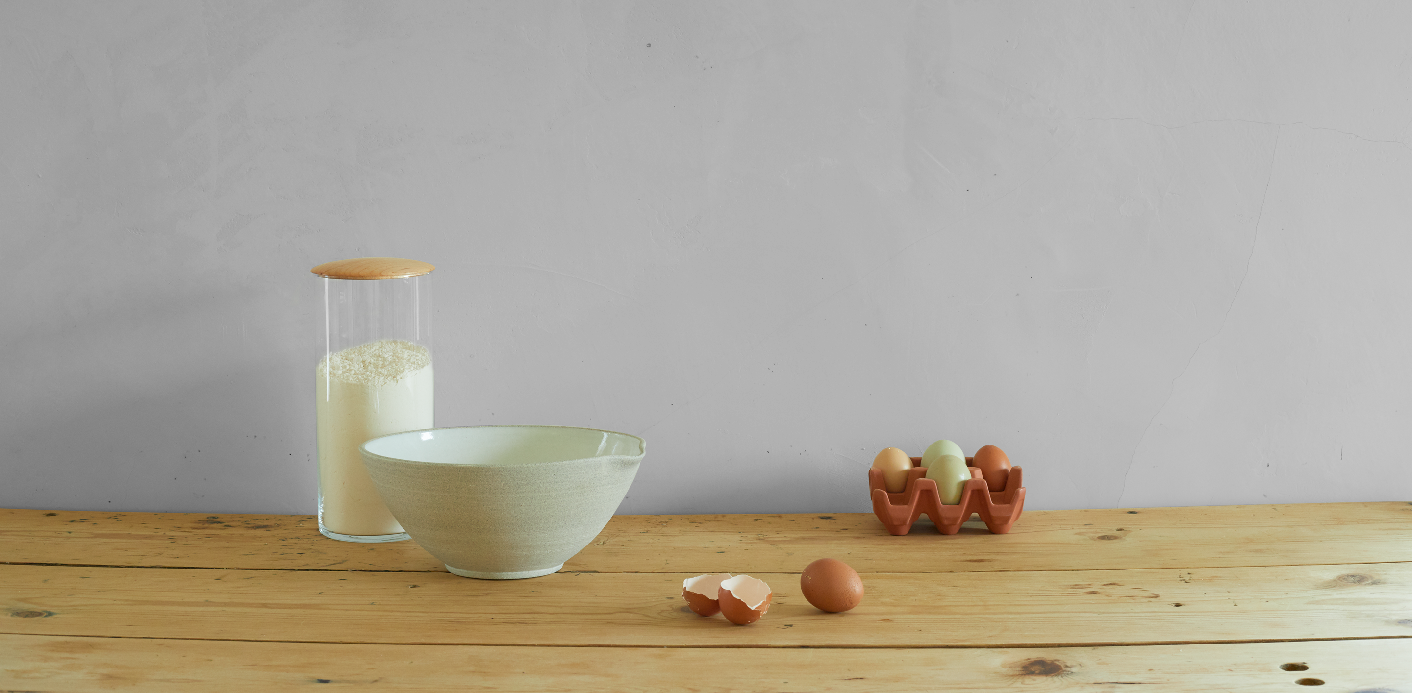 A ceramic bowl, a glass jar with flour, and a ceramic egg crate holding four eggs sit on a wooden surface. Two cracked eggshells are in front of the bowl.