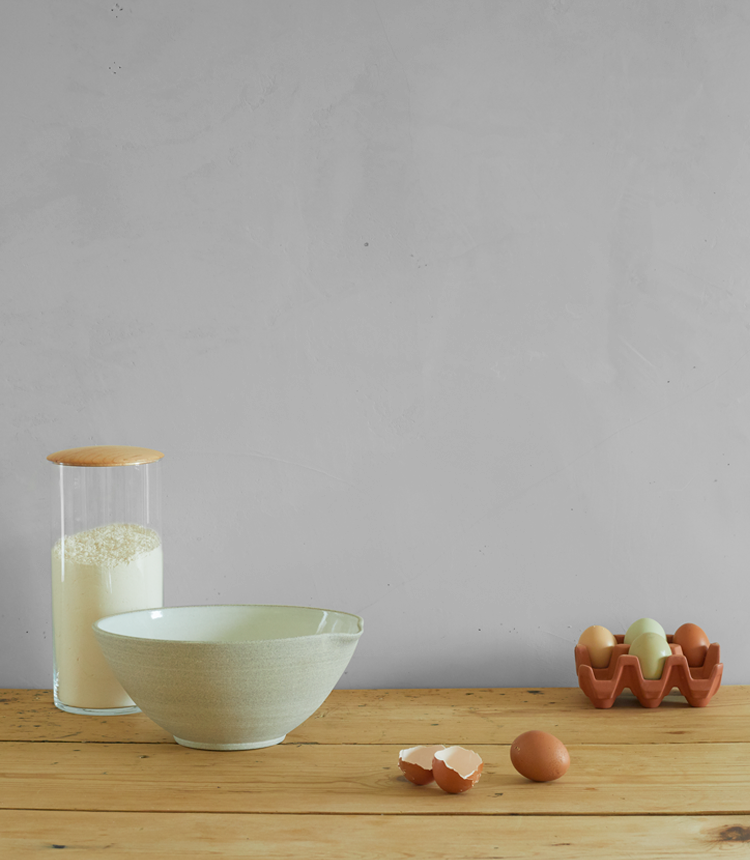 A ceramic bowl, a glass jar with flour, and a ceramic egg crate holding four eggs sit on a wooden surface. Two cracked eggshells are in front of the bowl.