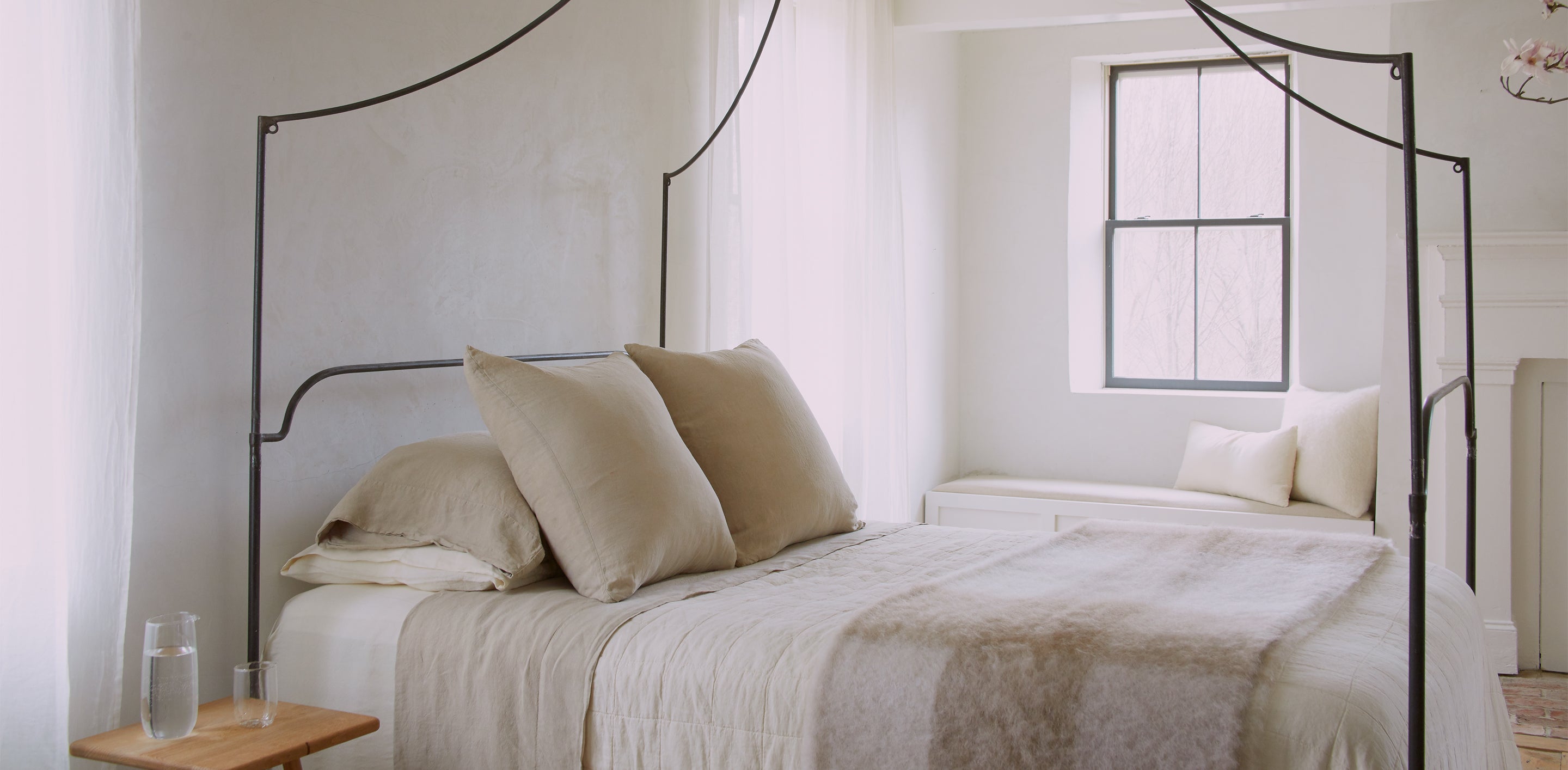 A minimalist bedroom features a bed with beige linens, a canopy frame, a wooden side table with a glass of water, a window with a cushioned bench, and white walls.