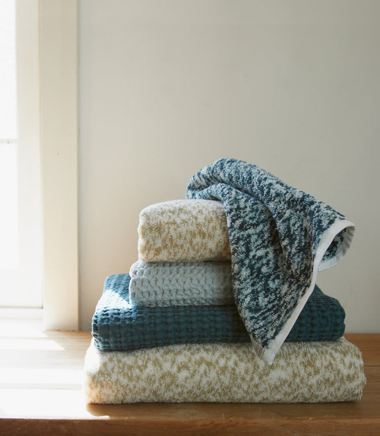 A stack of folded towels in various textures and shades of blue, gray, and beige sits on a wooden surface near a window.