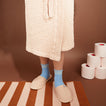 Person wearing blue socks and blush slippers and bathrobe on a striped bath mat with toilet paper rolls in the background.