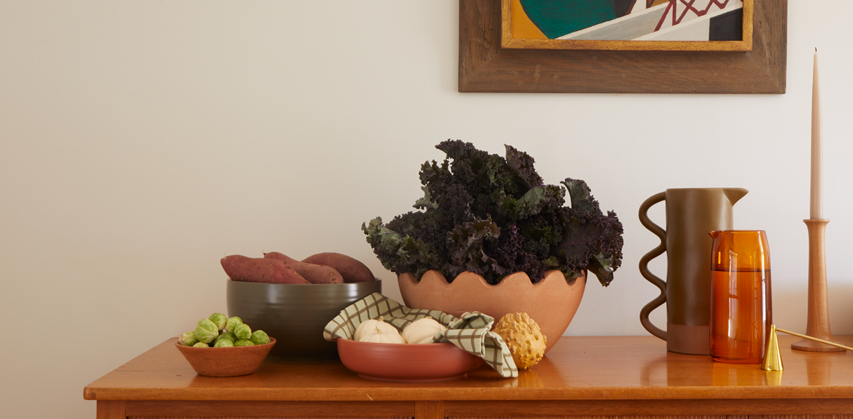 A dining table with serving bowls of various vegetables, to the right sits a clay and glass pitchers, a candle douter, taper candle, and artwork hangs in the background