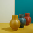 Essential ceramic vase in mustard, peacock, and terracotta on a yellow surface with a teal wall background.