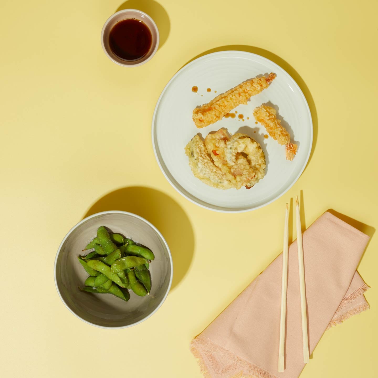 Plated tempura with edamame and chopsticks on a yellow background with xs bowl in blush and blush napkin.