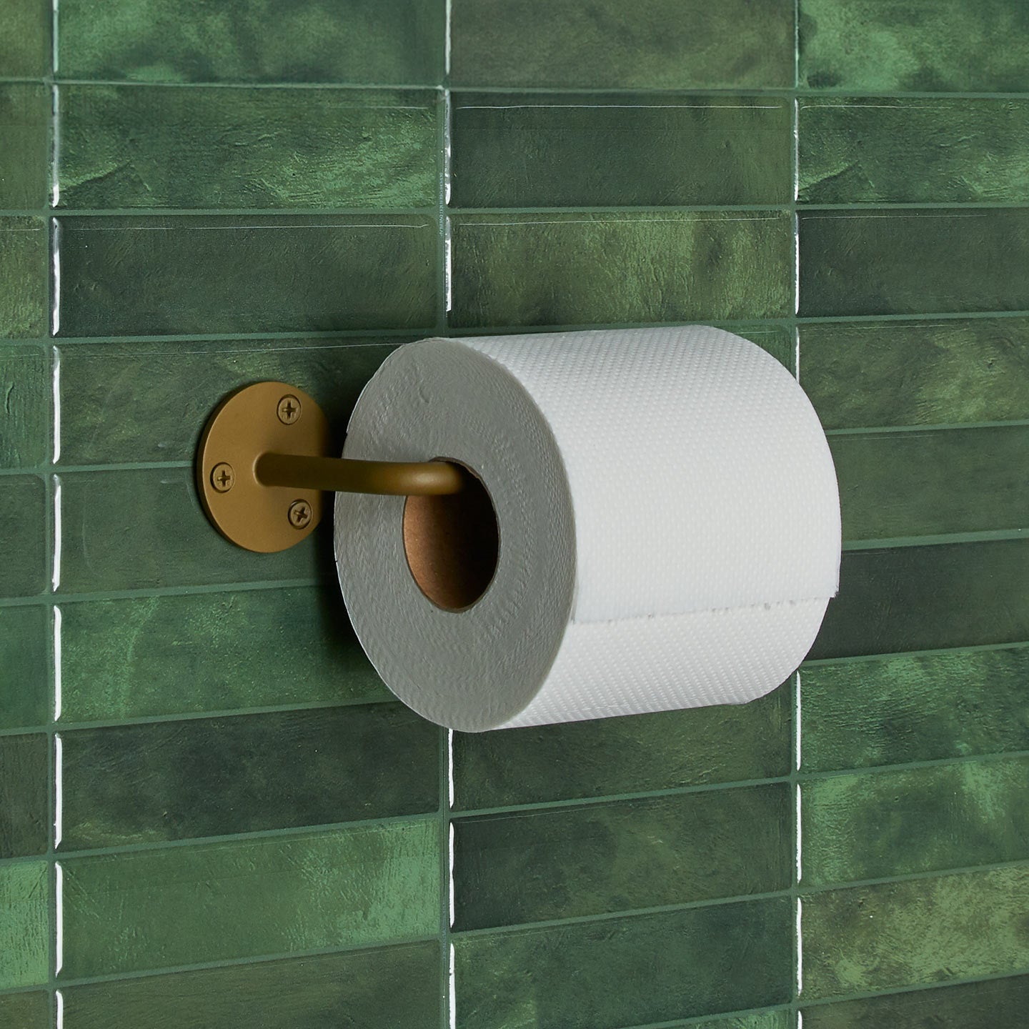 Essential toilet paper holder in Olive with a roll of toilet paper on tiled wall.