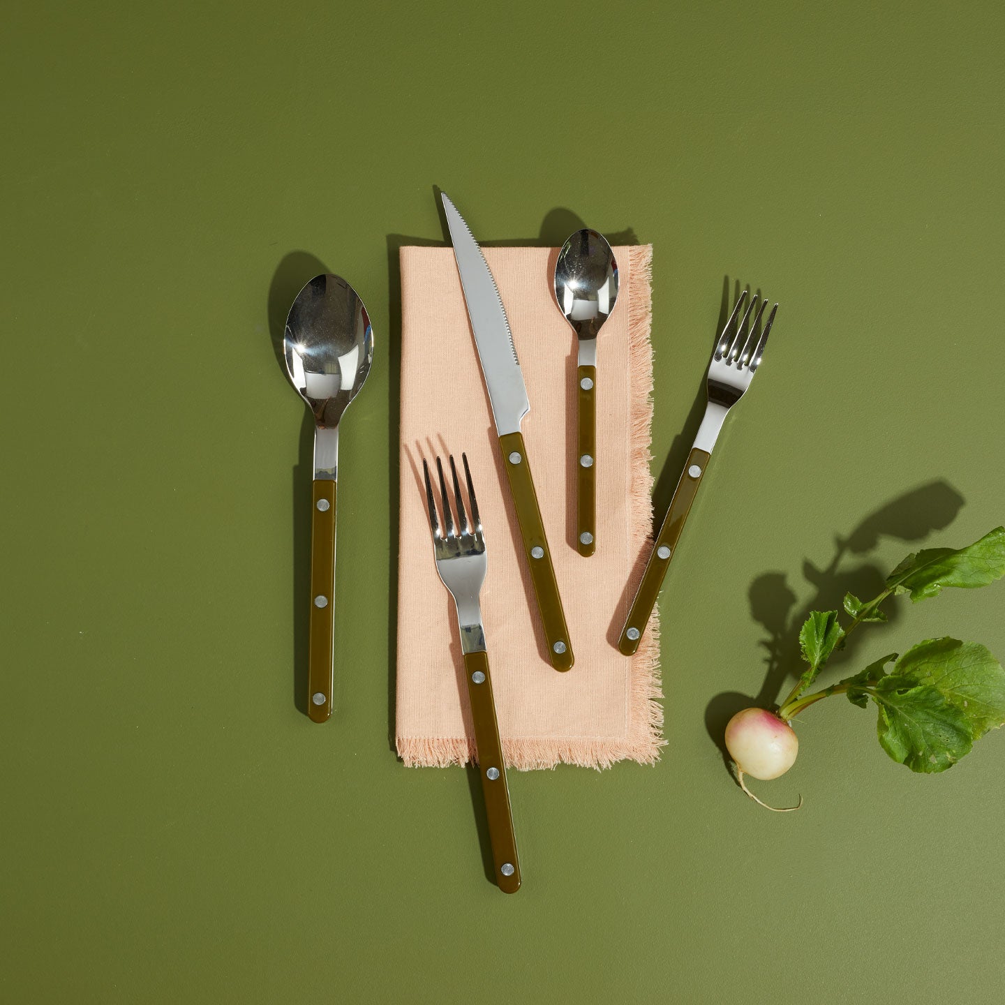 Olive flatware on folded Essential Napkin in Blush on green background with turnip.