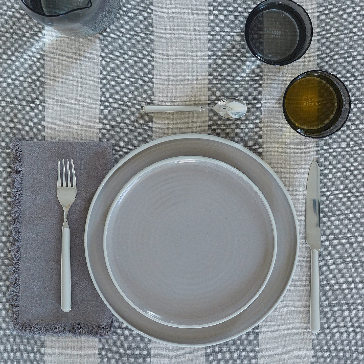 Placesetting with Essential Dinner Napkin in Dark Grey on grey striped tablecloth.