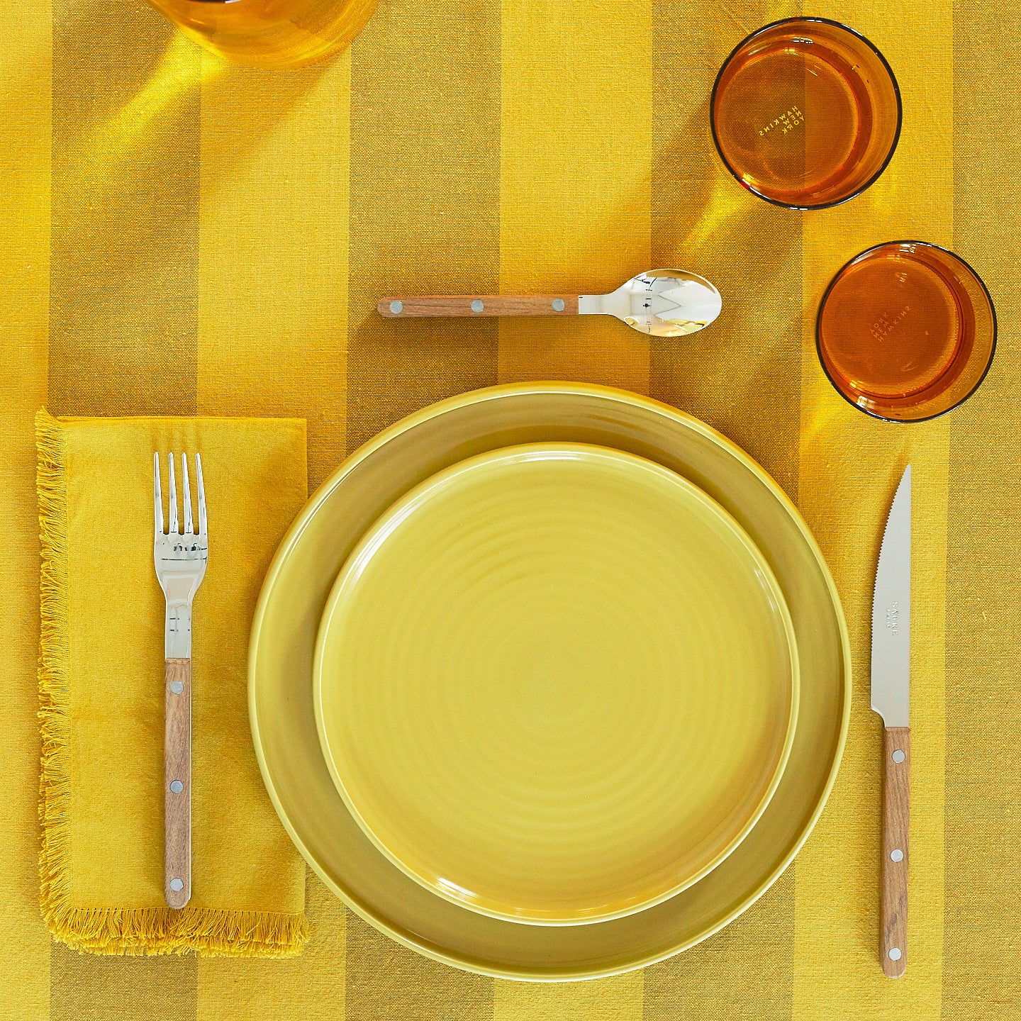 Placesetting with Essential Dinner Napkin in Mustard on yellow striped tablecloth.