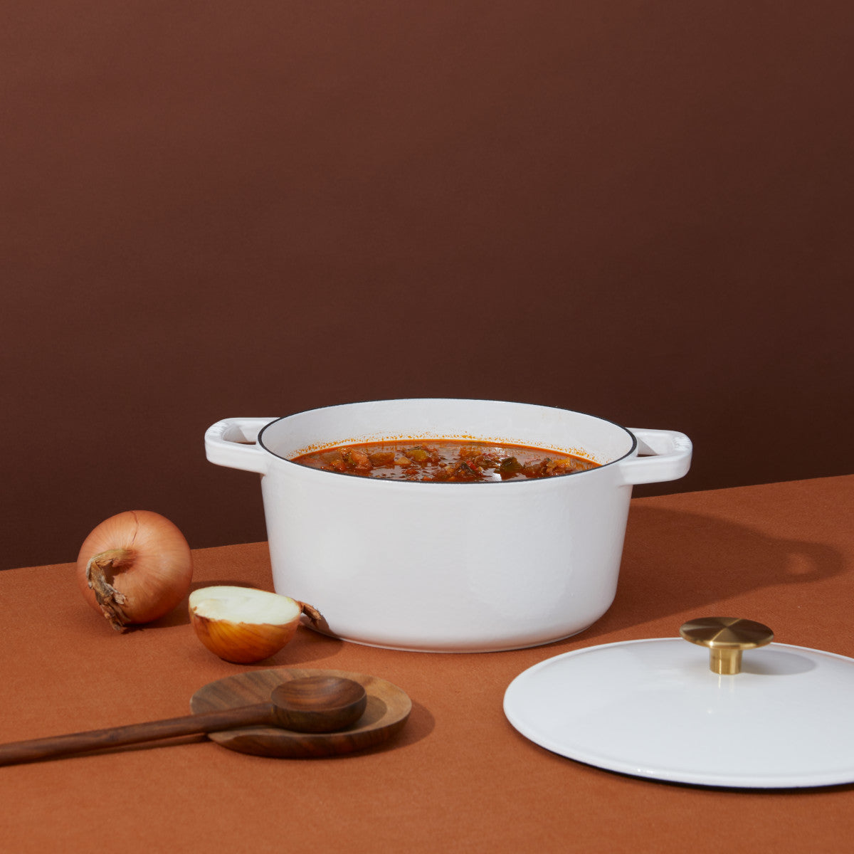 A white pot filled with soup sits on a brown surface, accompanied by a halved and a whole onion, a wooden spoon, and the pot's lid with a brass handle. Brown backdrop in the background.