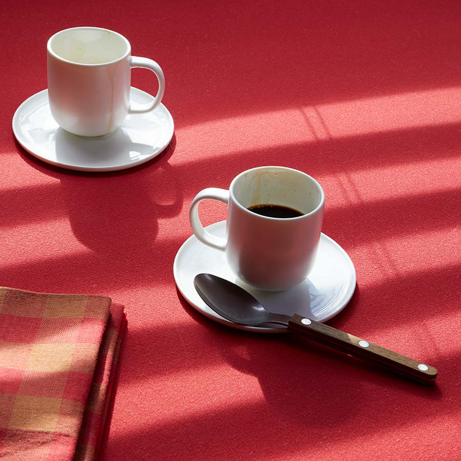 Two white coffee cups on a red tablecloth with a spoon and napkin.