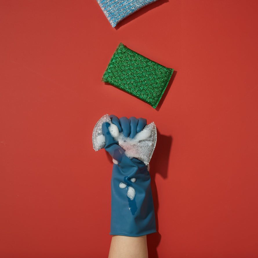 Hand wearing rubber glove squeezing soapy silver sponge on red background with green and blue sponges.