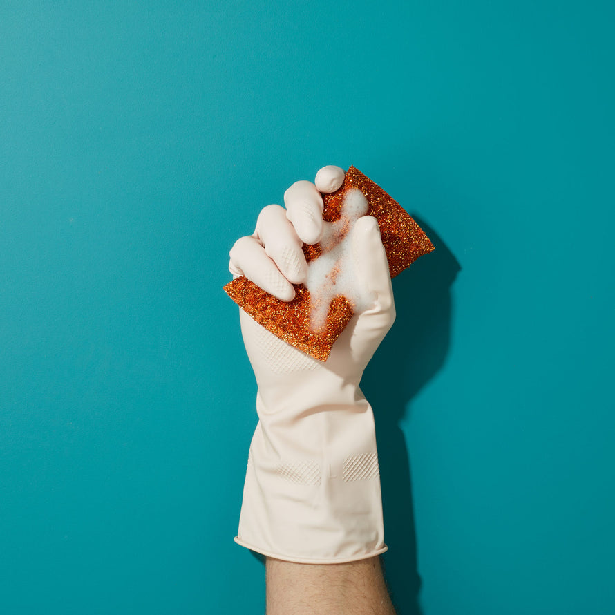 Hand wearing rubber glove squeezing soapy red sponge on blue background.