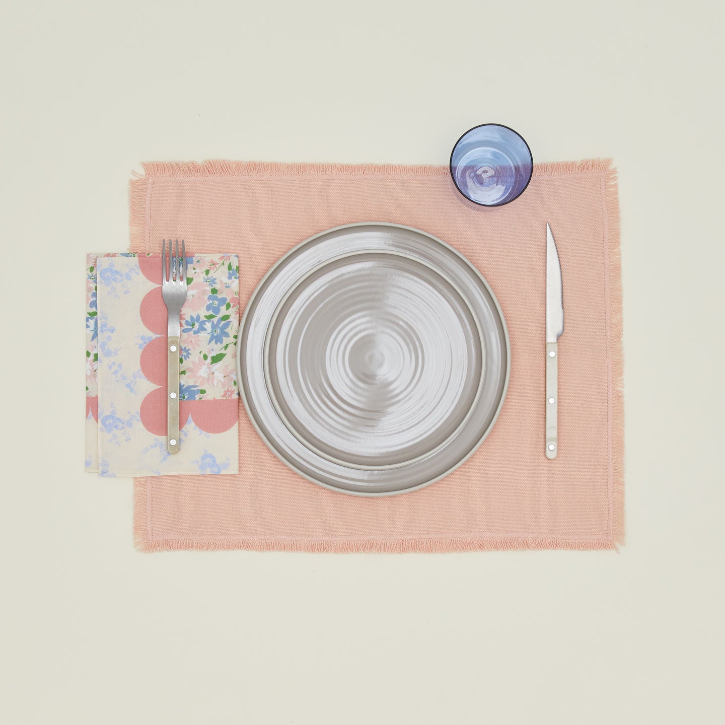 A table setting with grey plates, a blue glass, a blush placemat, and a rose patterned napkin.