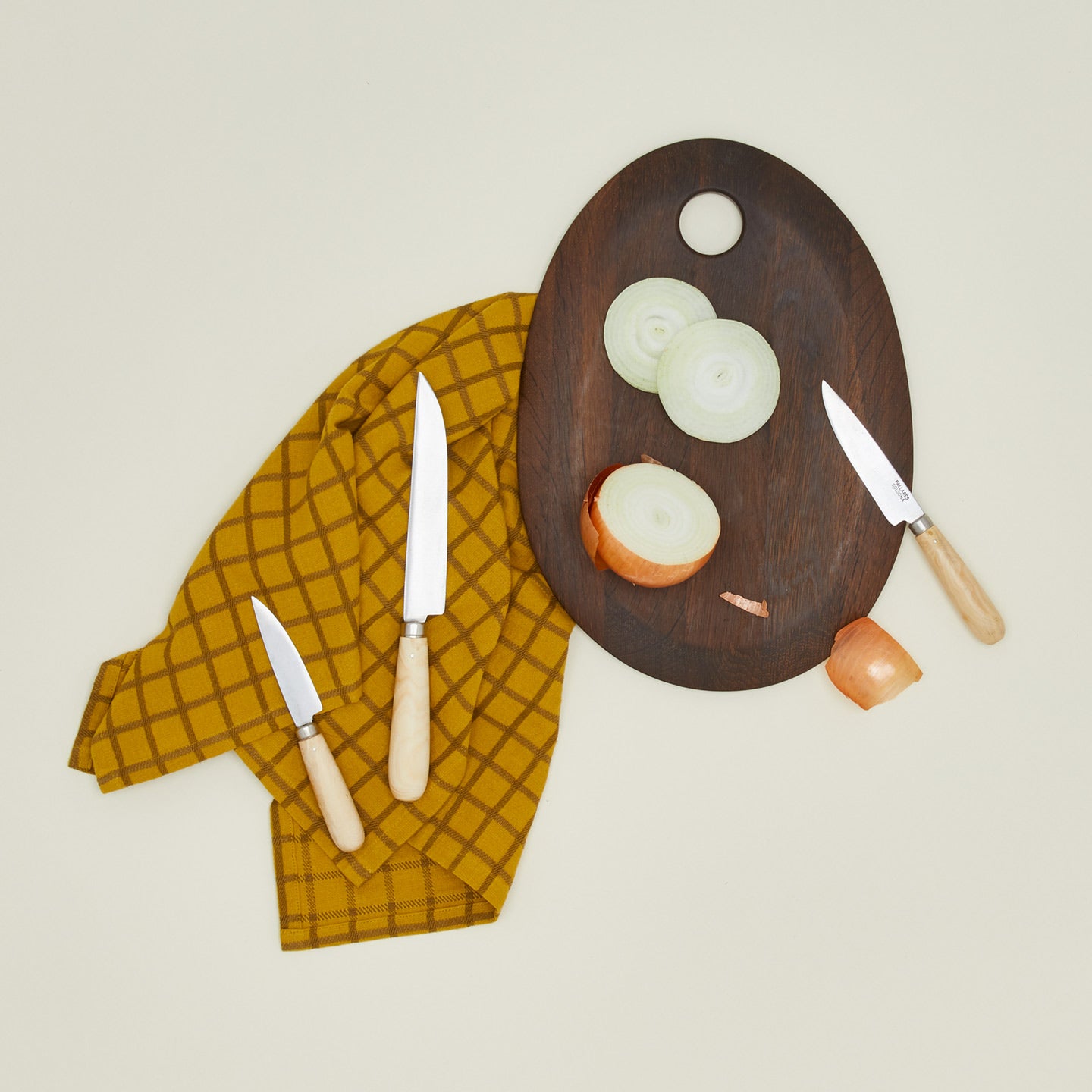 An overhead of three knives, a walnut cutting board, and a mustard dish towel.