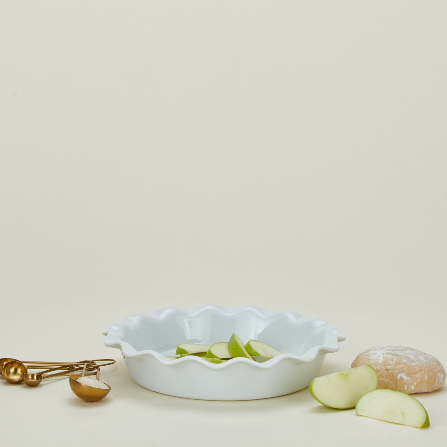 Porcelain pie dish in white with measuring spoons, dough, and apple slices