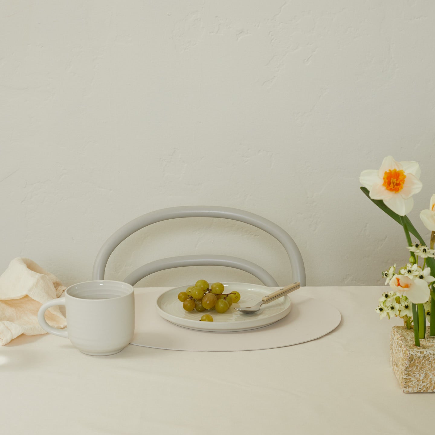 White mug on a table with a plate of grapes, a vase of flowers, and a chair in the background.