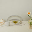 White mug on a table with a plate of grapes, a vase of flowers, and a chair in the background.